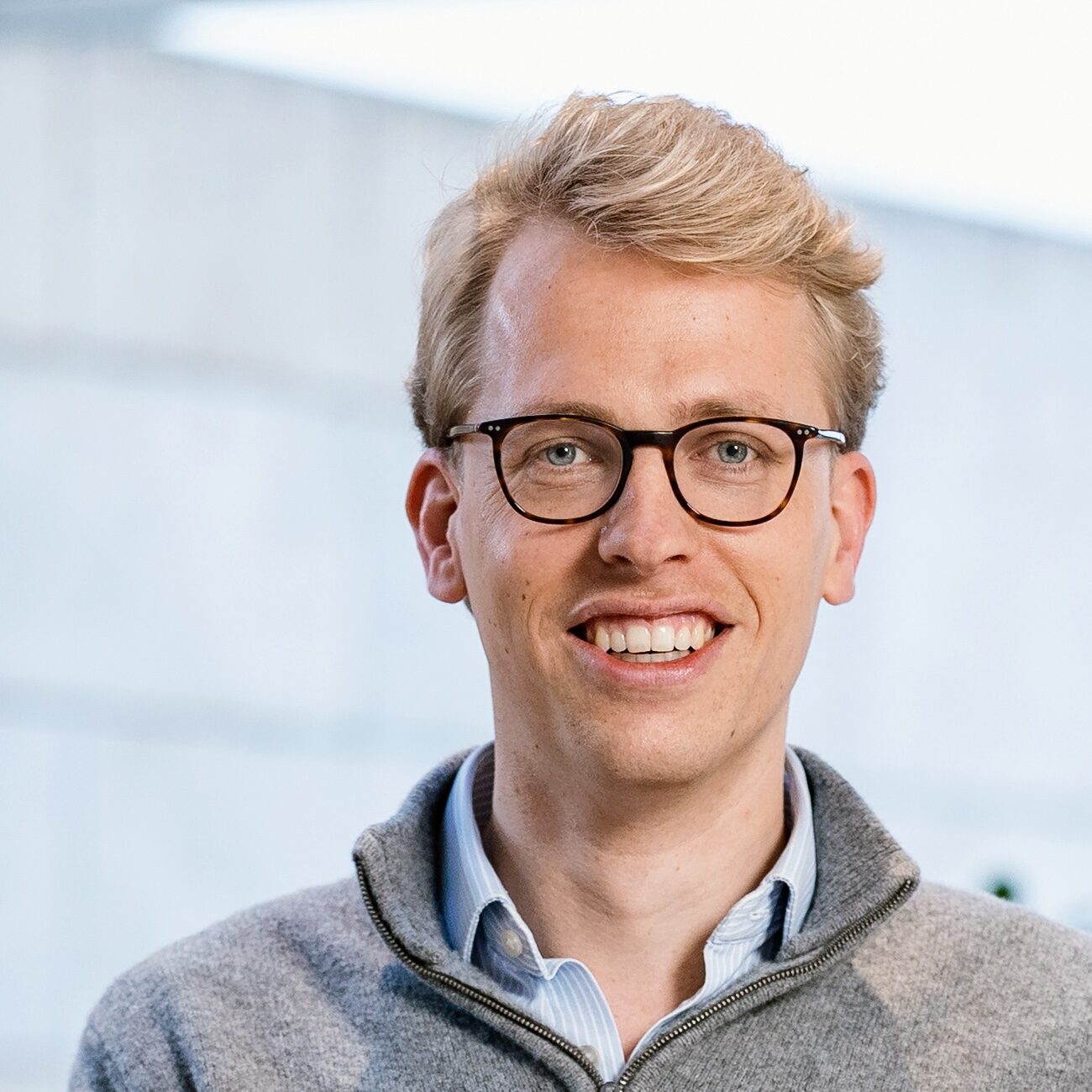 A young man with blond hair and glasses smiles at the camera. He is wearing a grey zip-up jumper over a collared shirt, with a blurred outdoor background.