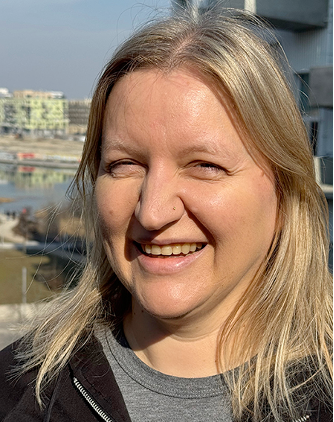 A woman with blonde hair smiles outdoors on a sunny day. Modern buildings and a body of water are visible in the blurred background.