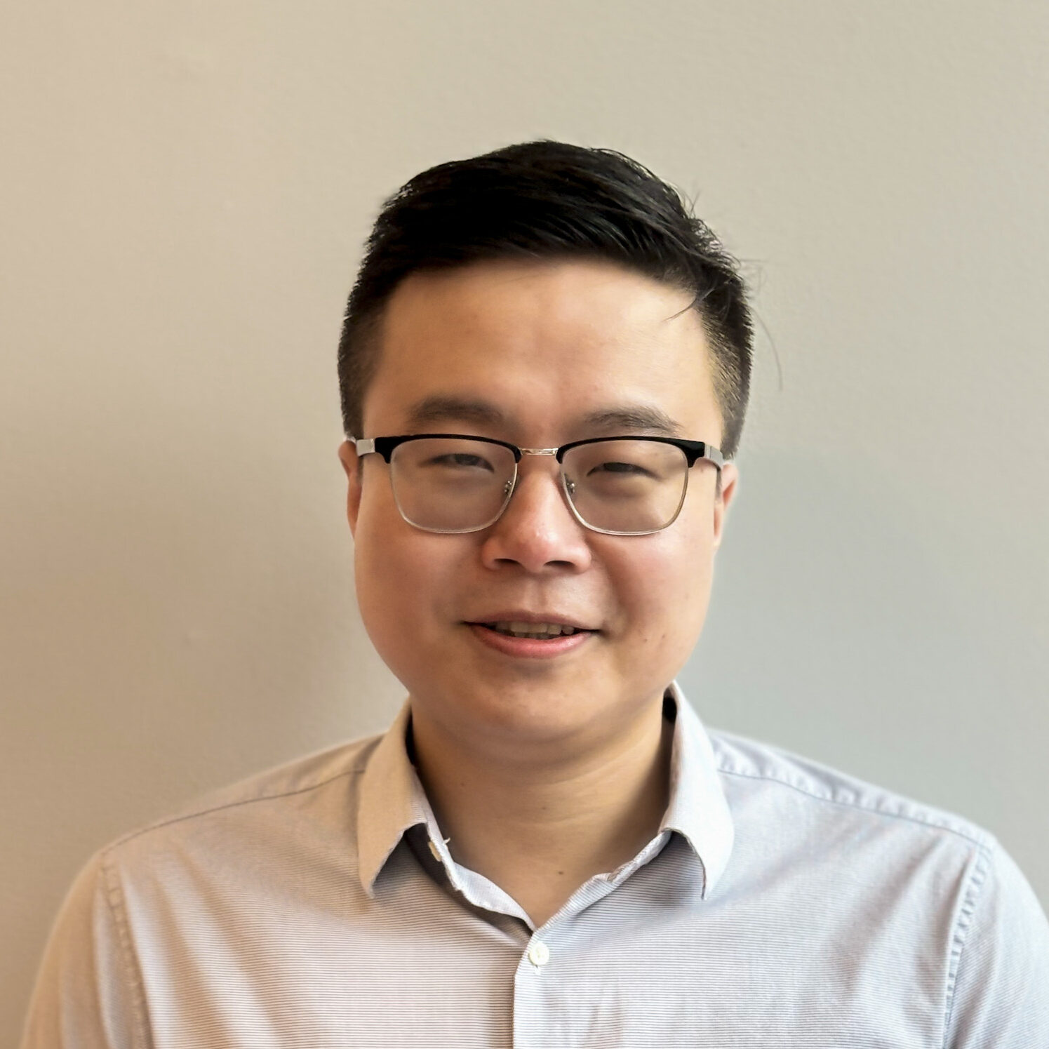 A person with short dark hair and glasses is smiling slightly, wearing a light grey button-up shirt, standing against a plain light-coloured wall.