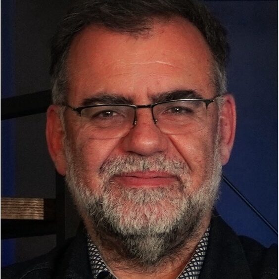 A middle-aged man with grey hair, a beard, and glasses is smiling slightly. He is wearing a dark jacket over a patterned shirt, standing indoors against a dark background.