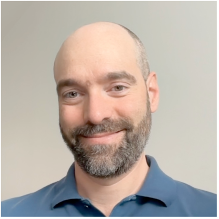 A man with a bald head and a short, neatly trimmed beard smiles gently at the camera. He is wearing a blue collared shirt and is posed against a plain, light-coloured background.