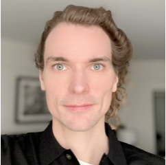A person with fair skin, light blue eyes, and wavy light brown hair poses indoors, wearing a black collared shirt. The background is softly focused, showing a framed picture and bright, neutral walls.