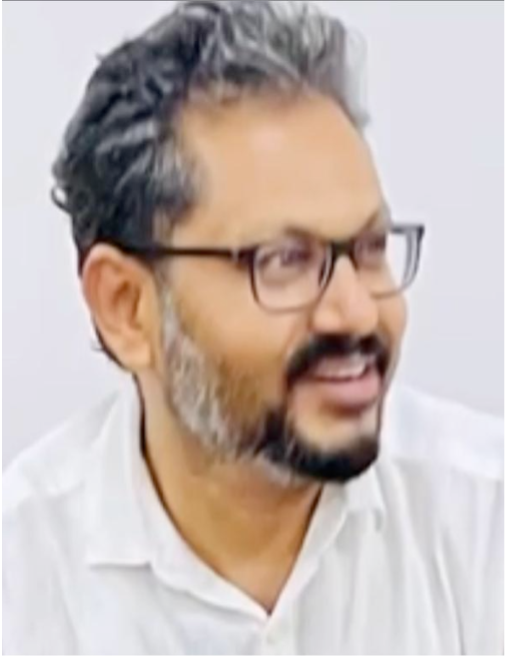A man with short, curly dark hair and a beard, wearing glasses and a white shirt, smiling whilst looking to the side against a plain light background.