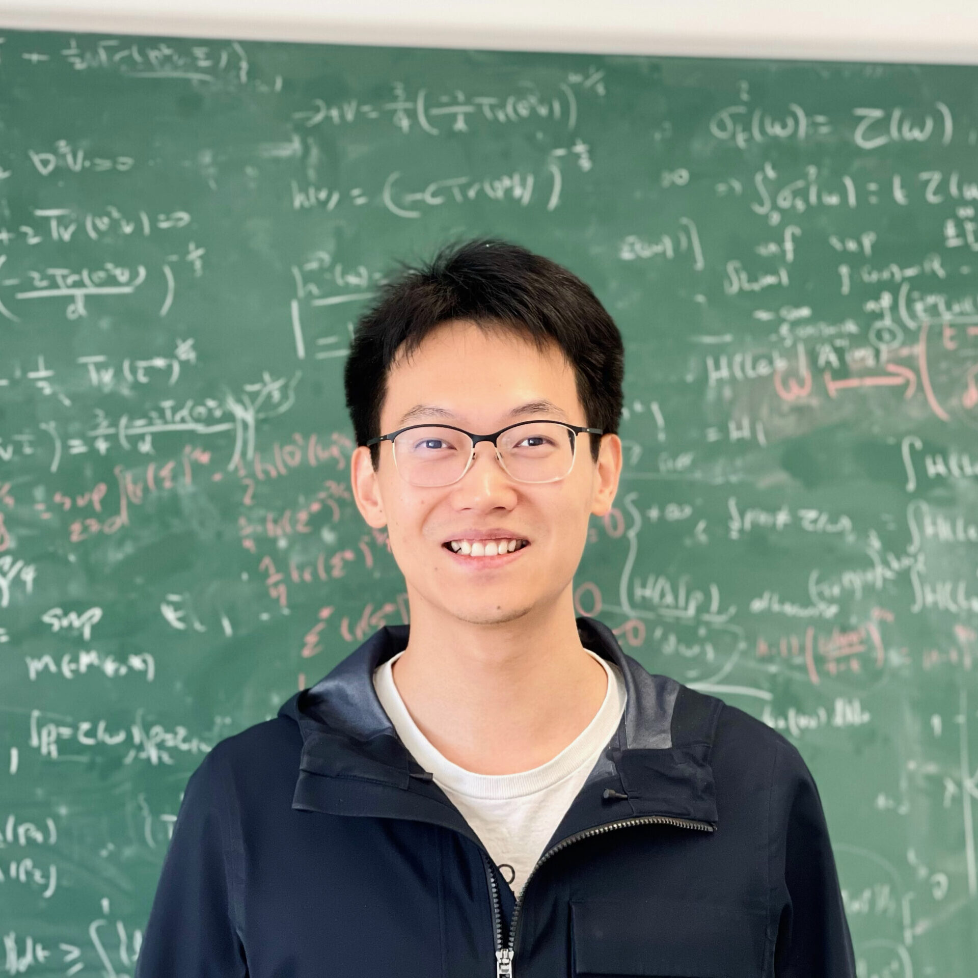 A person wearing glasses and a dark jacket is smiling in front of a green blackboard filled with complex mathematical equations written in white chalk.