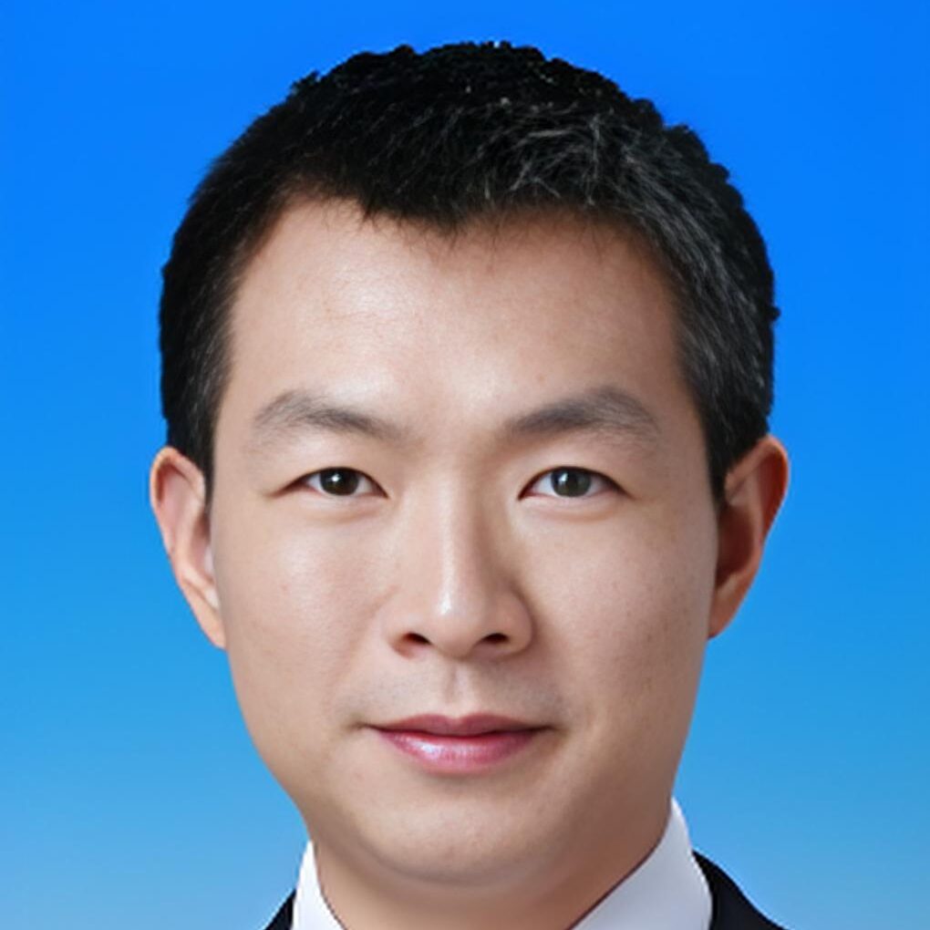 A man with short black hair wearing a black suit, white shirt, and striped tie poses against a blue studio background.