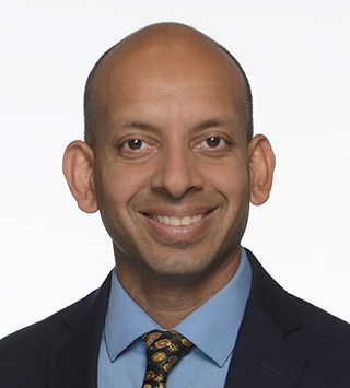 A smiling man with a shaved head, wearing a dark suit, blue shirt, and patterned tie, posing against a plain white background.