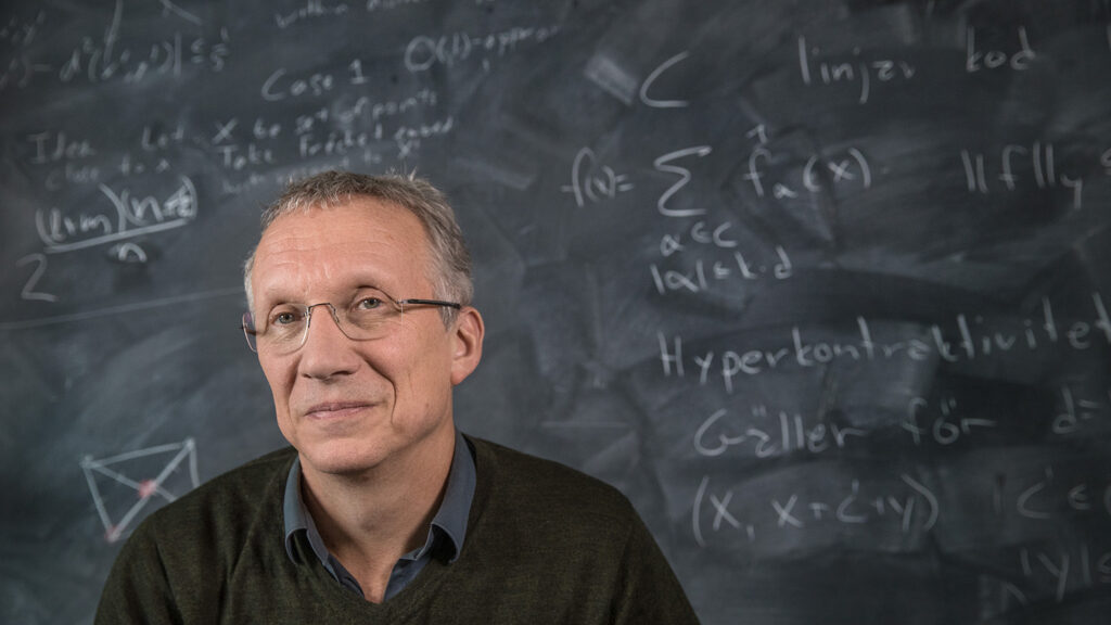 A man with glasses and grey hair sits in front of a blackboard covered with mathematical equations and symbols, looking slightly to the side and smiling gently.