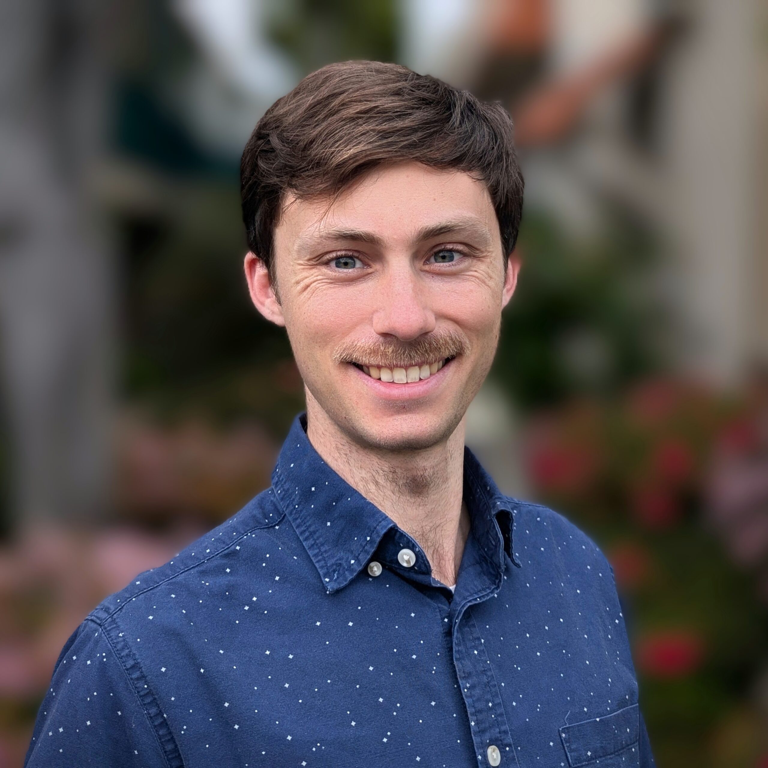 A young man with light skin, short brown hair, and a moustache smiles at the camera. He wears a blue, button-up shirt with small white dots. The background is blurry with greenery and colourful flowers.