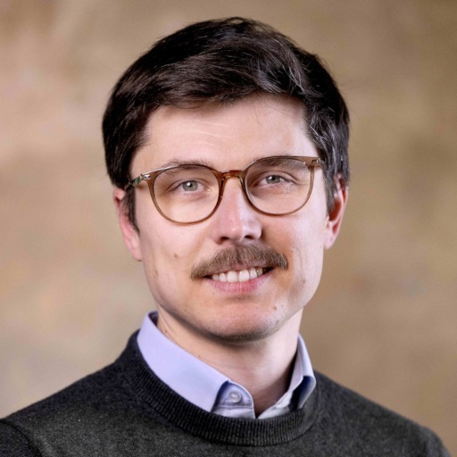 A man with short brown hair and a moustache, wearing round glasses, a blue collared shirt, and a dark jumper, smiles in front of a neutral beige background.
