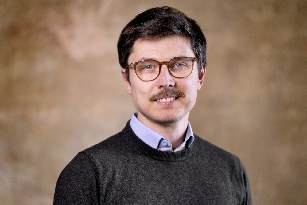 A man with short brown hair, glasses, and a moustache is wearing a dark jumper over a collared shirt. He is smiling slightly and standing in front of a brown, softly blurred background.