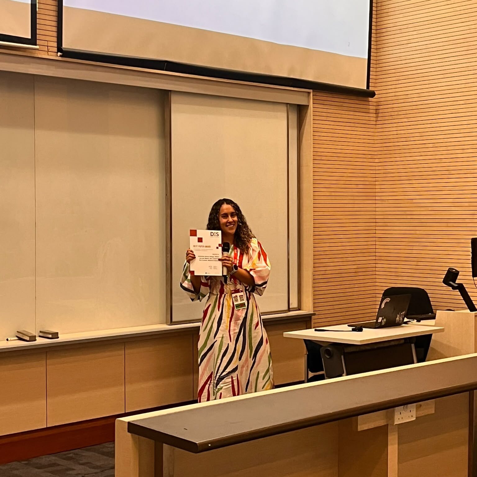 A woman in a colourful dress stands at the front of a lecture theatre holding a certificate. Behind her is a large projector screen displaying a yellow warning message. A lectern and laptop are visible nearby.