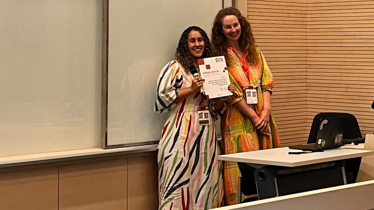 Two women stand in a conference room, both smiling at the camera. One is holding a certificate. They are wearing colourful dresses and conference badges, next to a desk and whiteboard.