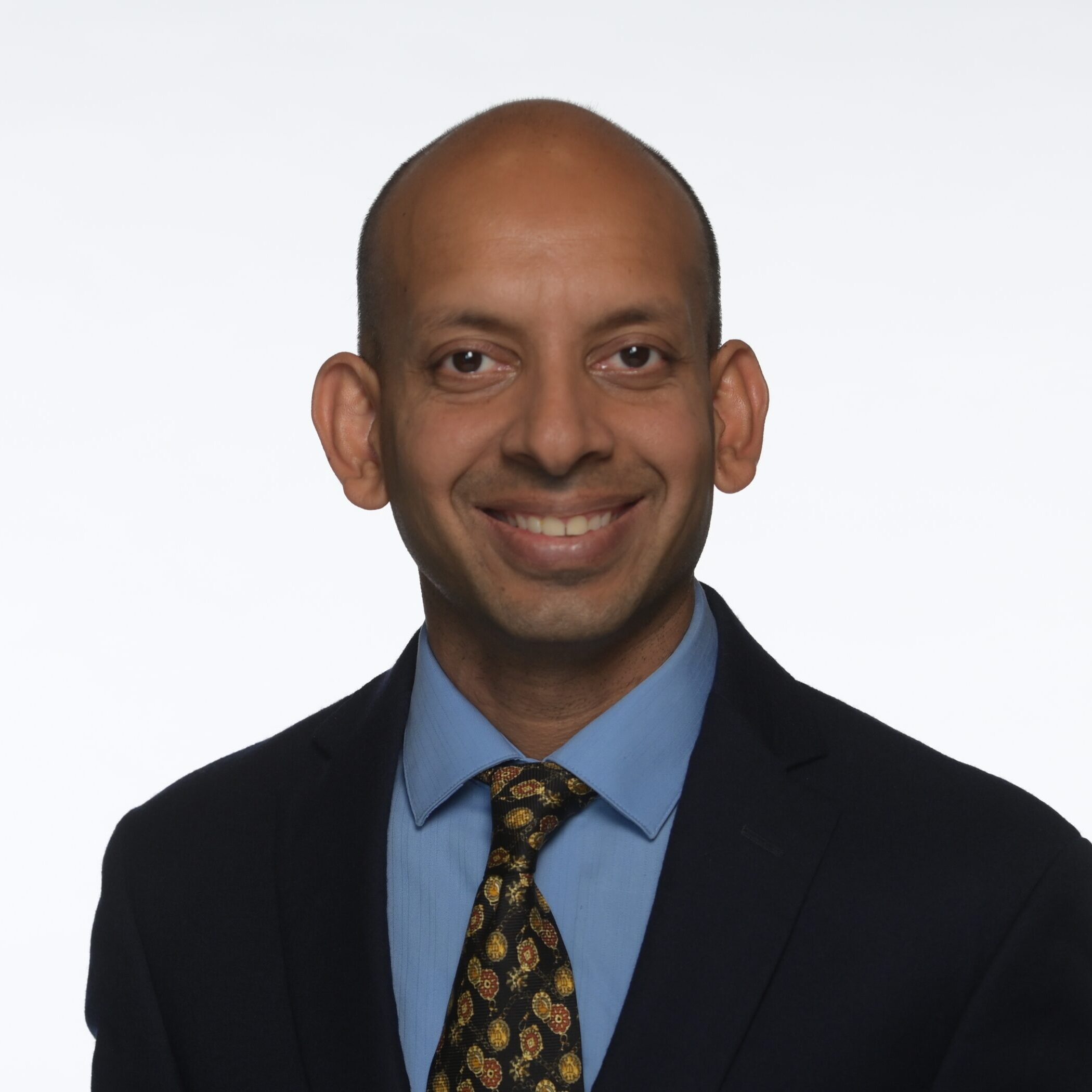A smiling man with a bald head wears a dark suit, blue shirt, and patterned tie, posing against a plain white background.