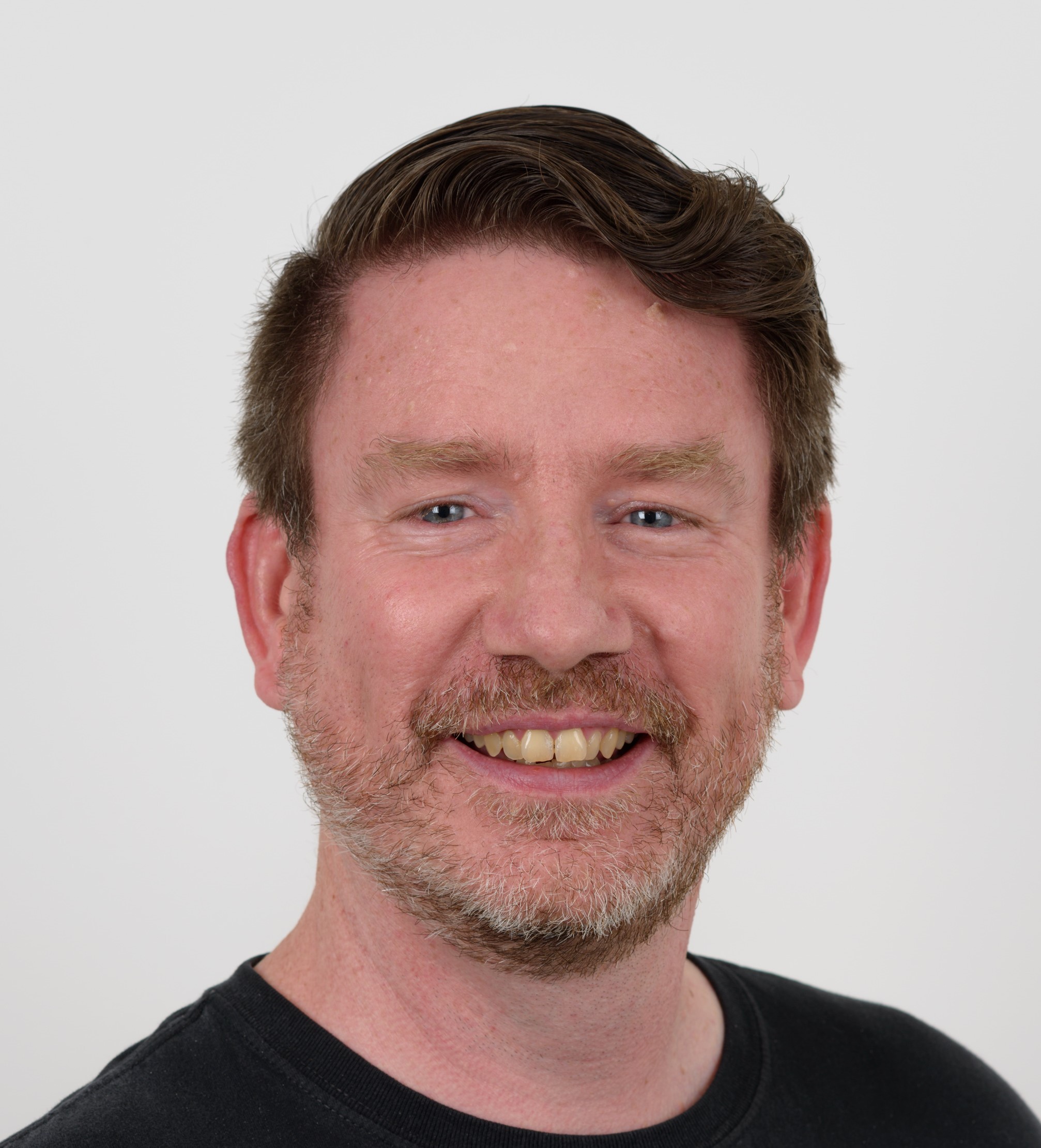 A man with short brown hair and a beard smiles at the camera against a plain white background. He is wearing a black shirt.