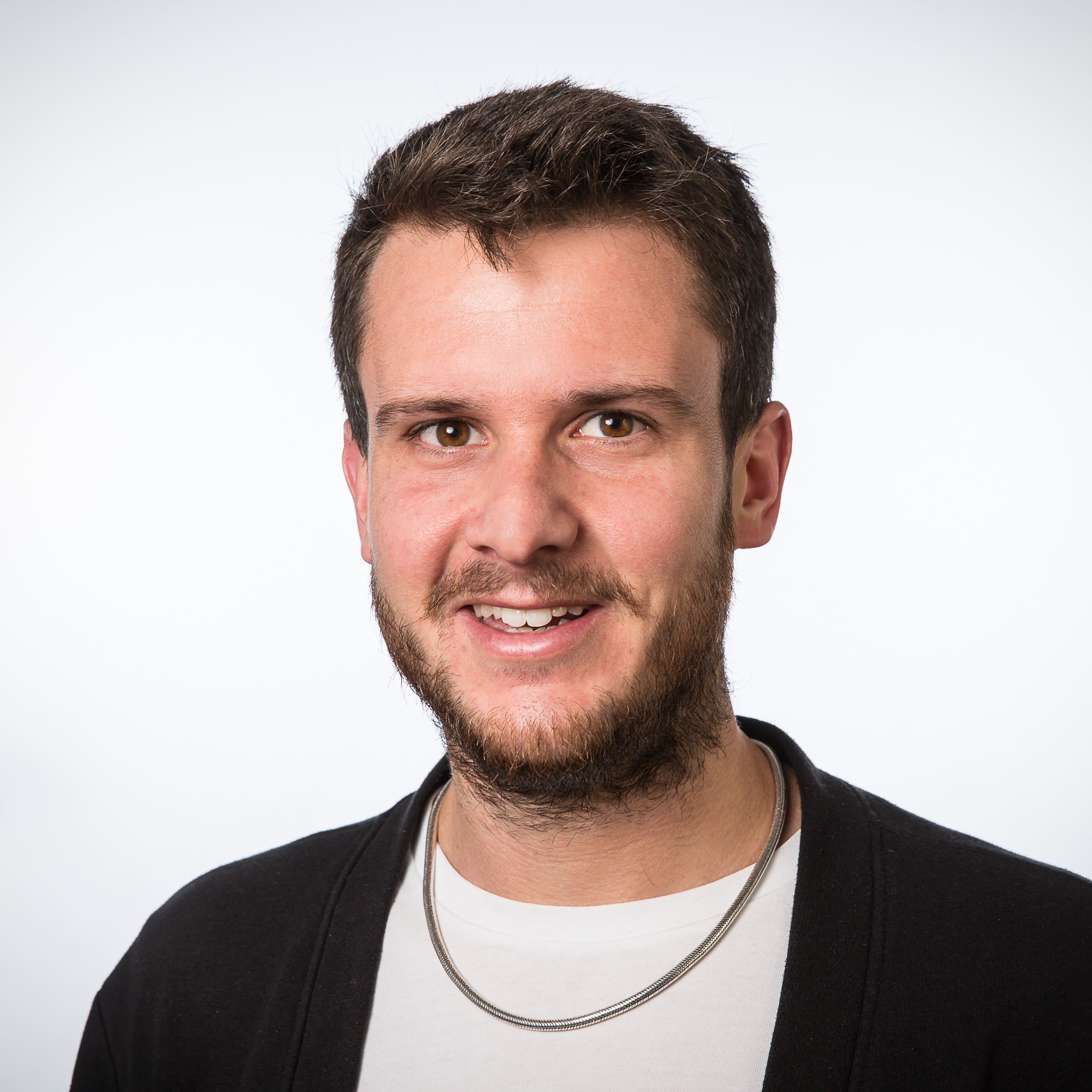 A man with short brown hair and a beard smiles at the camera. He is wearing a white shirt, a dark cardigan, and a silver necklace, with a plain white background behind him.