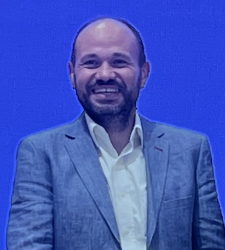 A man with a beard and short hair, wearing a light grey blazer over a white shirt, stands and smiles in front of a solid bright blue background.