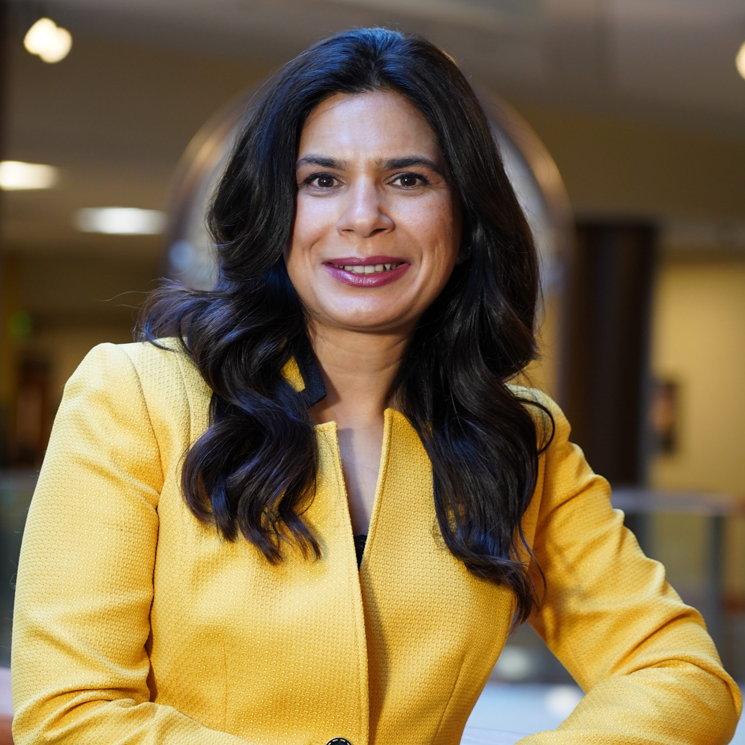 A woman with long dark hair wearing a bright yellow blazer smiles confidently at the camera in an indoor setting with soft background lighting.