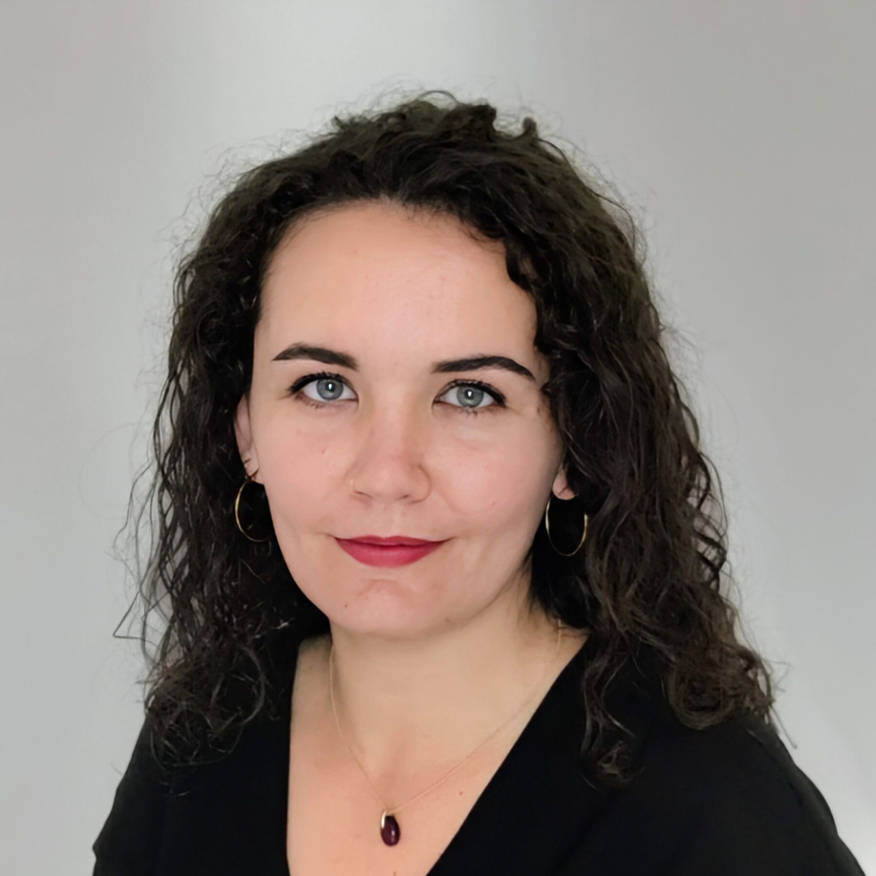A woman with curly dark hair, wearing hoop earrings, a black blouse, and a pendant necklace, poses in front of a plain light grey background. She is looking at the camera with a slight smile.