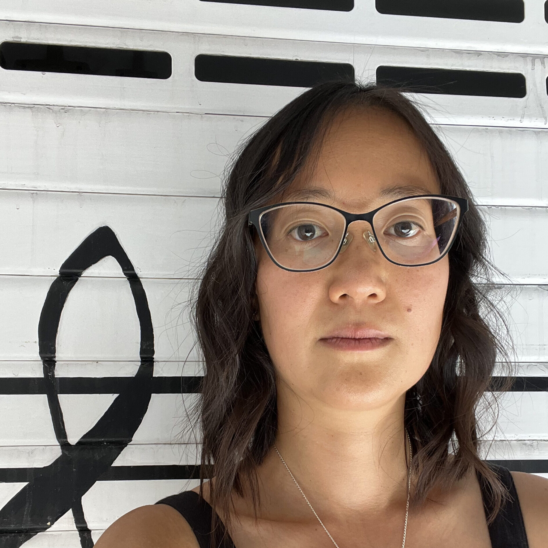 A woman with shoulder-length wavy hair and glasses stands in front of a white wall with a large black treble clef symbol. She wears a black top and a silver necklace, looking directly at the camera.