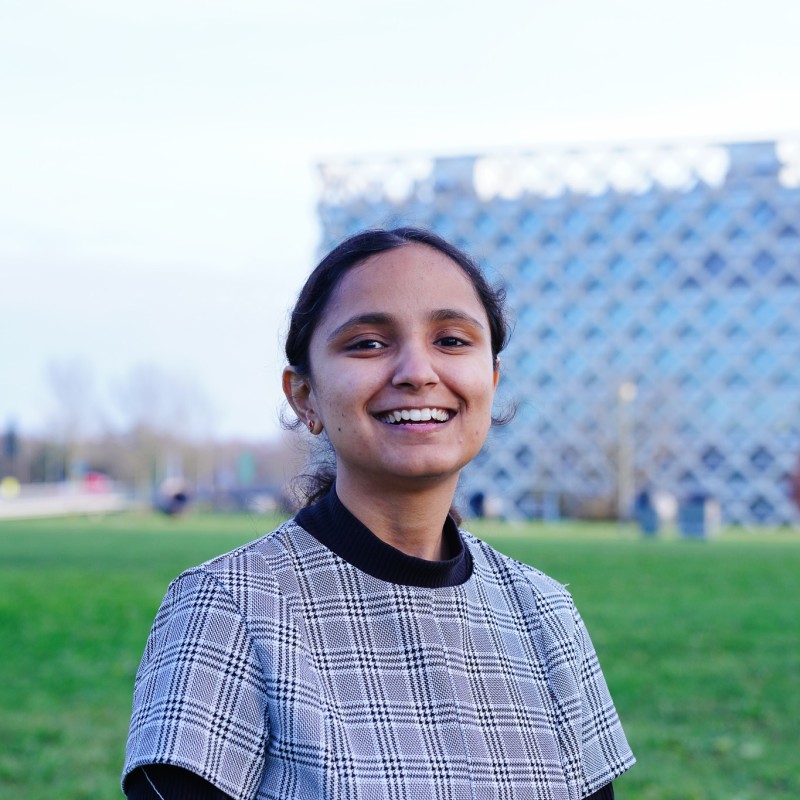 A young woman with dark hair tied back, wearing a grey checked top, smiles at the camera whilst standing on a green lawn with a modern, patterned building in the background.