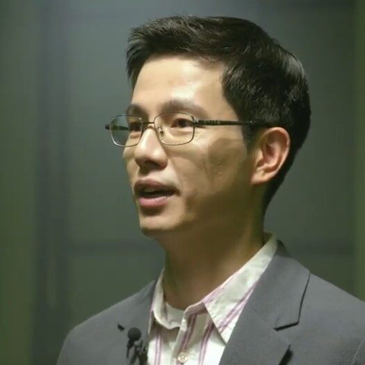 A man with short dark hair and glasses, wearing a grey blazer and a light-coloured collared shirt, speaks while facing slightly to the side. A microphone is clipped to his jacket. The background is out of focus.