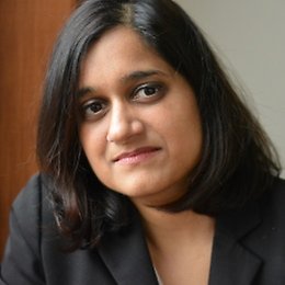 A person with straight, shoulder-length black hair is wearing a black blazer and looking at the camera with a neutral expression. The background is softly blurred.