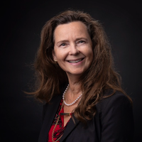A woman with long brown hair wearing a dark blazer, red patterned blouse, and a pearl necklace smiles against a dark background.