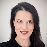 A woman with long dark hair and fair skin smiles at the camera. She is wearing a black top and has red lipstick. The background is plain and white.