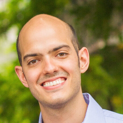 A man with a shaved head and a light beard is smiling at the camera. He is wearing a light blue button-up shirt and standing outdoors with greenery in the blurred background.