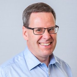 A man with short brown hair, wearing glasses and a light blue collared shirt, smiles at the camera against a plain light grey background.