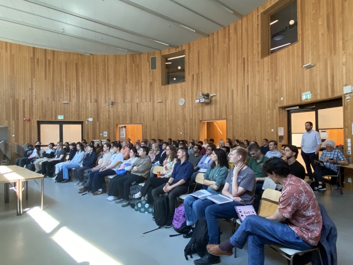 A group of people sits in rows of chairs in a large, modern lecture theatre with wooden walls, attentively listening to a presentation. Some people hold notebooks or laptops. A presenter stands by the door on the right.