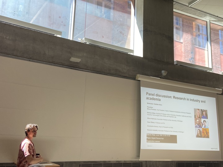 A person stands at a lectern in a bright room with large windows, presenting a slide titled Panel discussion: Research in industry and academia that lists panellists and their photos.