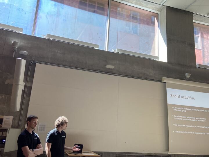 Two people stand in front of a projector screen giving a presentation in a modern room with large windows and concrete walls. The screen displays a slide titled Social activities.
