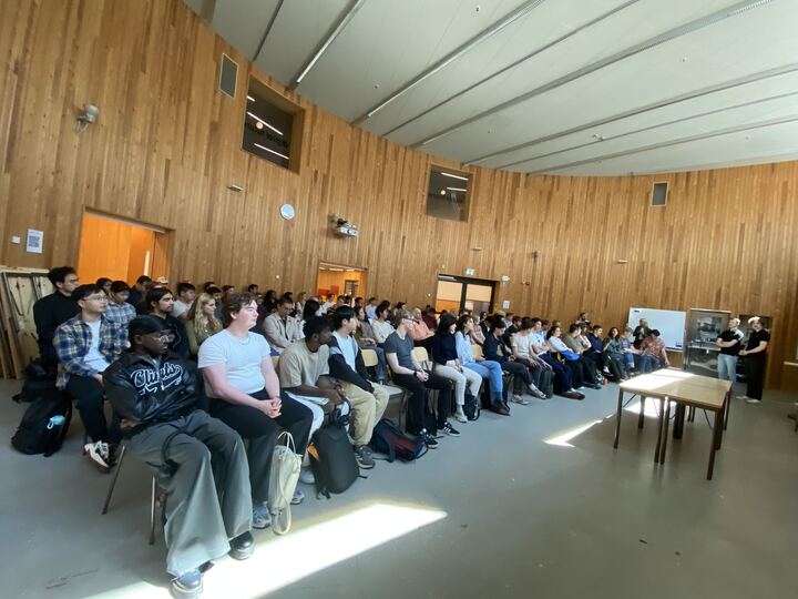 A large group of people sits attentively in rows of chairs in a spacious room with wooden walls and a high curved ceiling, whilst a few individuals stand and speak at the front near a table.