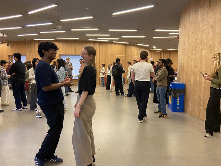 People stand and chat in a spacious, modern room with wooden walls. Most are in casual clothes, and some hold drinks. A blue recycling bin is visible. The atmosphere appears social and relaxed.