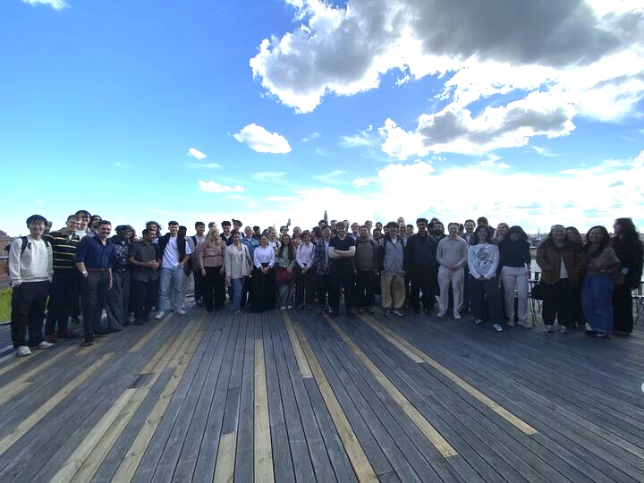 A large group of people stand together on a wooden decking outdoors under a partly cloudy blue sky, posing for a group photo. Most people are facing the camera, with buildings visible in the distant background.