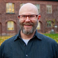 A smiling, bearded man wearing glasses and a dark striped shirt stands outdoors in front of a brick building with large windows.