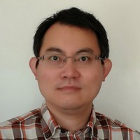 A man with short dark hair and glasses wearing a checked shirt faces the camera in front of a plain light-coloured background.