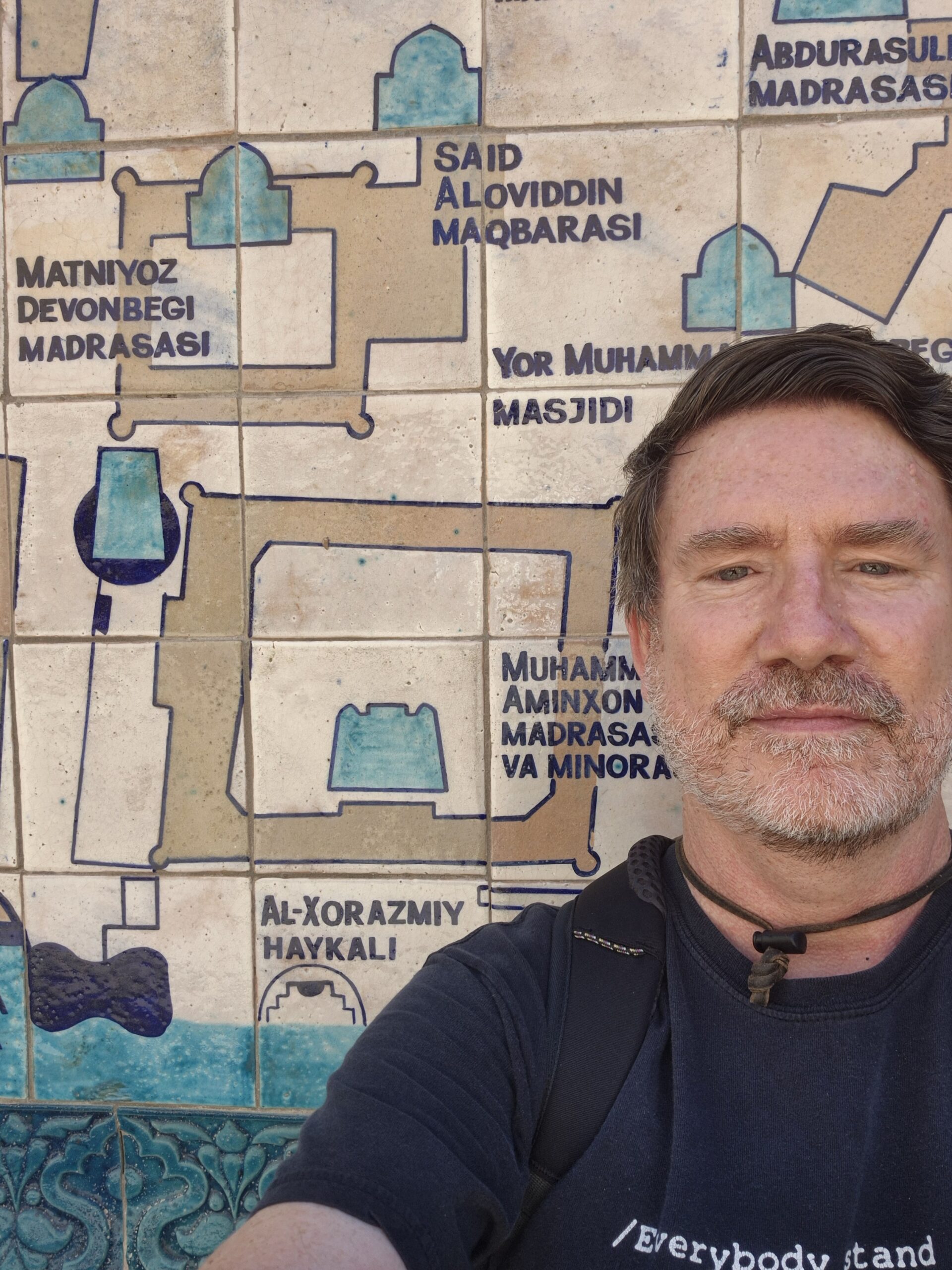 A man with short brown hair and a beard takes a selfie in front of a tiled wall map featuring sites and names written in Uzbek and English, including madrasahs and mosques.