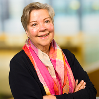 An older woman with short grey hair, wearing a black top and a colourful pink and orange scarf, smiles with her arms crossed. The background is softly blurred with light and yellow tones.