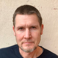 A man with short brown hair and a trimmed beard is looking at the camera. He is wearing a dark shirt and standing in front of a light-coloured textured wall.