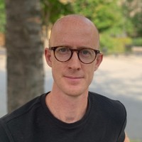 A bald man with glasses and a black shirt is sitting outdoors. There are blurred green trees and a tree trunk in the background.