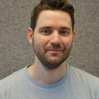 A man with short brown hair and a beard, wearing a light grey T-shirt, smiling softly at the camera in front of a textured grey background.