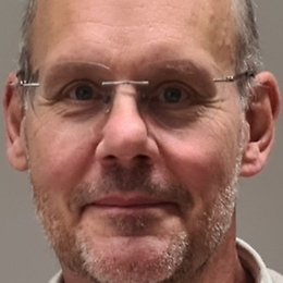 A middle-aged man with short hair, wearing glasses and a light-coloured shirt, looks directly at the camera with a neutral expression.