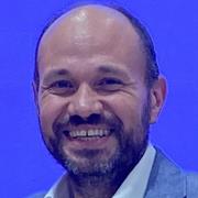 A man with a beard and moustache smiles at the camera. He is balding and wearing a light blue blazer. The background is a solid bright blue.