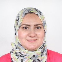 A woman wearing a patterned light-coloured hijab and a pink top, smiling at the camera against a plain white background.