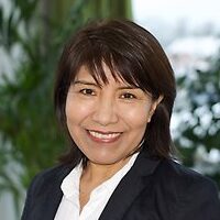 A woman with short dark hair, wearing a dark blazer and white shirt, smiles at the camera. She is indoors with green plants and a window with natural light in the background.