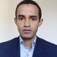 A man with short dark hair and brown eyes, wearing a navy blazer and light blue collared shirt, looks directly at the camera with a neutral expression against a plain light background.