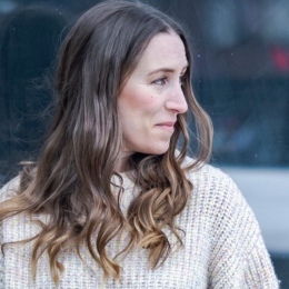 A woman with long, wavy brown hair and a cream-coloured jumper looks to her right, standing outdoors with a blurred background.