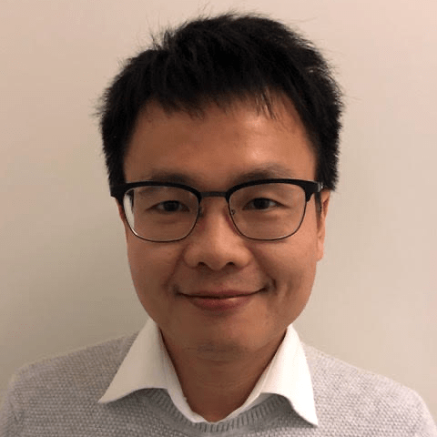A person with short black hair and glasses is smiling slightly. They are wearing a white collared shirt under a light grey jumper, standing in front of a plain light-coloured wall.